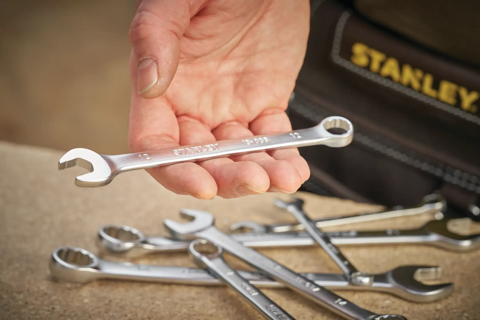 STANLEY FMMT13035-0 spanner held in hand, with other wrenches on a workbench, close-up angle.